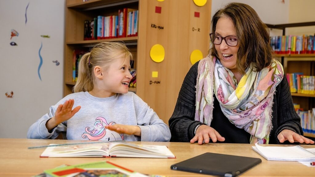 Foto von einem Mädchen und einer Frau, die gemseinsam an einem Tisch sitzen, sich ansehen und mit den flaschen Händen auf den Tisch klopfen
